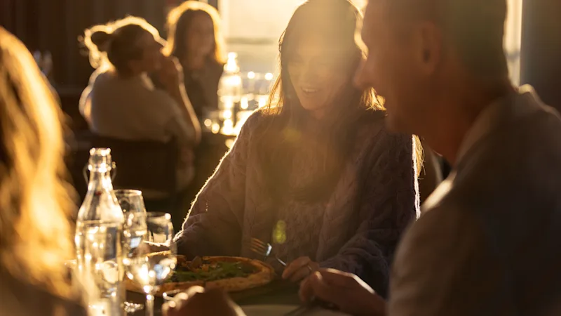 En kvinna njuter av pizza vid ett bord med andra i en varmt belyst restaurang. Solljus filtreras genom fönstret och skapar en mysig känsla vid middagsbordet.