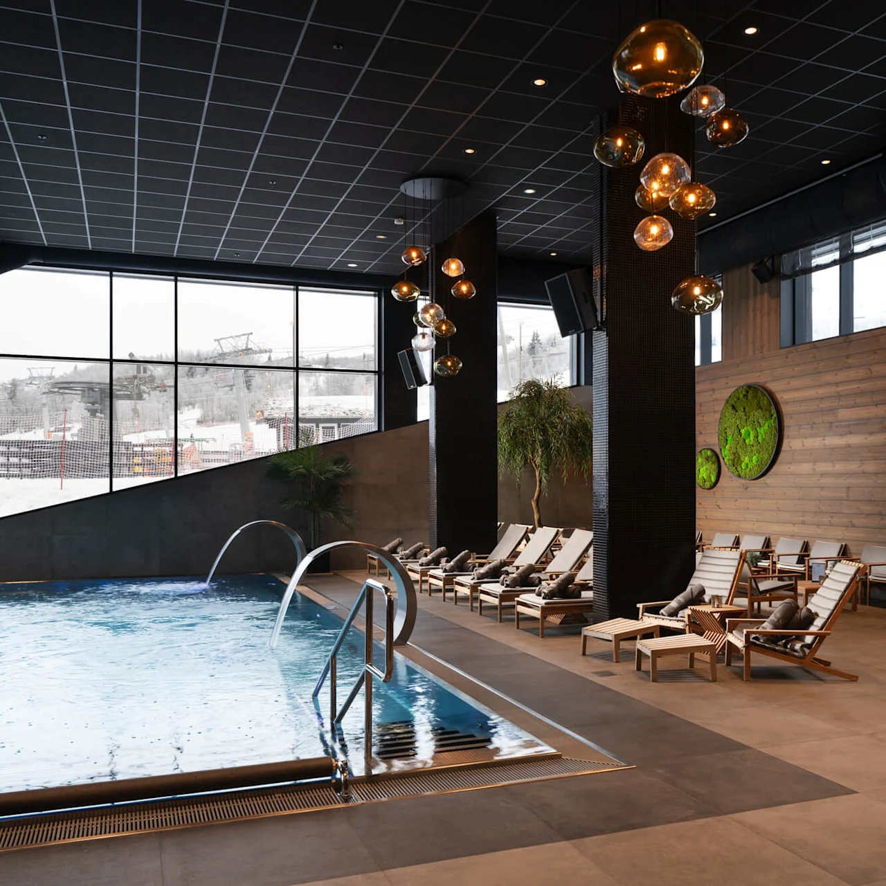 En swimmingpool med krusende vand, omgivet af liggestole, er inde i et moderne rum med store vinduer med udsigt over snedækkede bjerge. Hængelamper og planter fremhæver spa-lignende atmosfære.