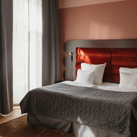 A neatly made bed with a grey quilt and white pillows is placed against a red headboard, near a green mini-fridge with snacks, in a softly lit room.