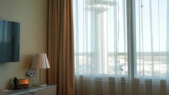 A room features a lamp and phone on a cabinet next to a large window with sheer curtains. Outside, a view of a tower against a clear sky is visible.