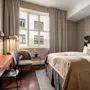 En pent oppredd seng med hvitt og beige sengetøy står i et ryddig rom. Nær en skrivebord, stol og lenestol ved et vindu med trukne gardiner.
