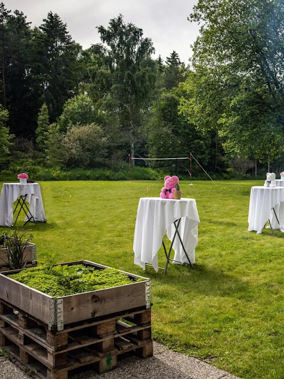 Runde bord med hvite duker viser rosa og hvite blomsteroppsatser. Et stort telt og trær gir en naturskjønn bakgrunn i en gresskledd utendørs arrangementsetting.
