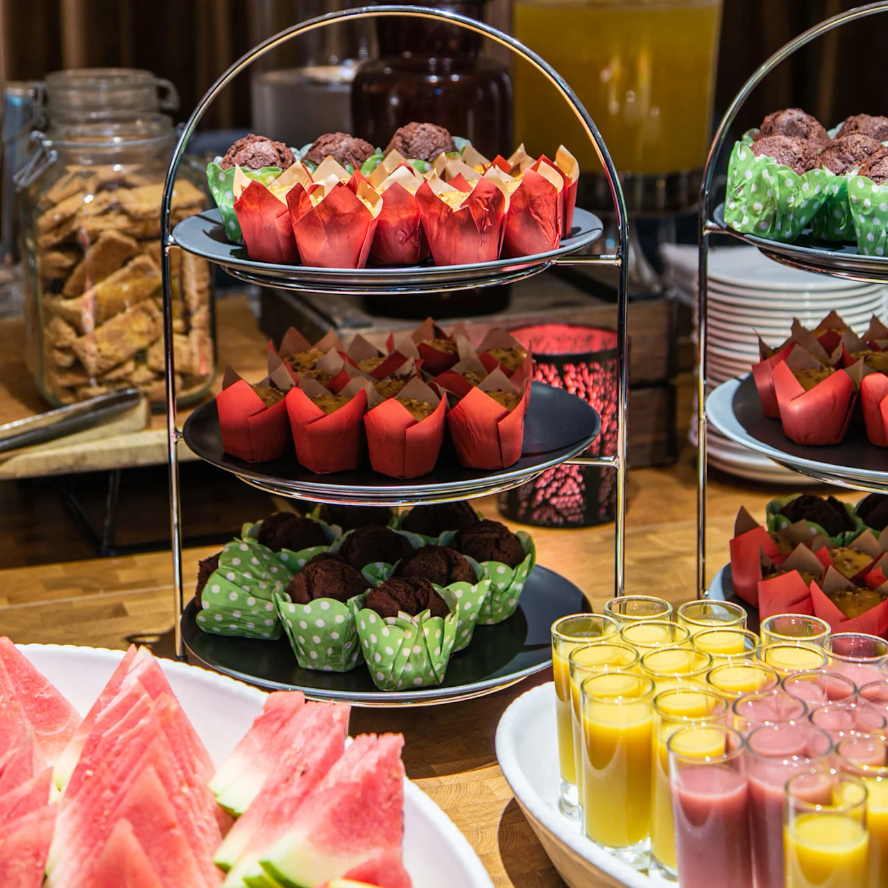 Cupcakes arrangeret på et stasionsstativ, omgivet af melonskiver og juiceglas, sat i en buffetopsætning med krukker med snacks og dekorative tørre grene i baggrunden.