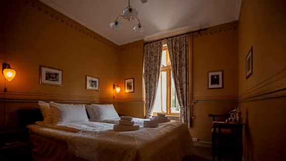 A bed with white linens and neatly folded towels is in a cozy, warmly lit room with beige walls, decorative wall lights, framed pictures, and a window with patterned curtains.