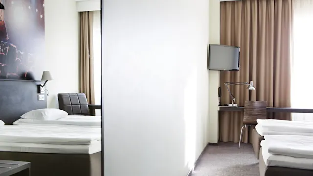Two neatly made beds are on opposite sides of a light-filled hotel room, separated by a partial wall. A desk with a lamp, chair, and TV is visible.