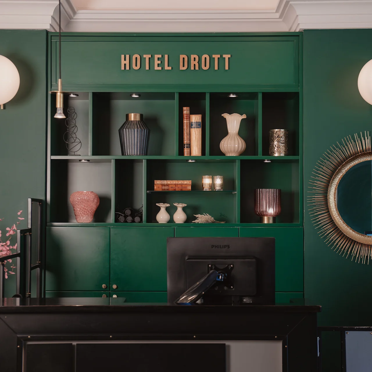 Reception med lyserøde blomster foran en grøn hylde med vaser og bøger. "HOTEL DROTT" er skrevet ovenover. Runde spejle og lamper pryder væggene.