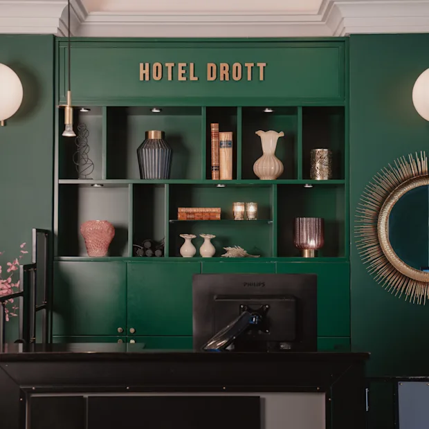 Reception med rosa blommor framför en grön hylla med vaser och böcker. "HOTEL DROTT" står skrivet ovanför. Runda speglar och lampor pryder väggarna.