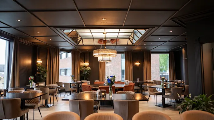 An elegant restaurant dining room with multiple tables set for guests, featuring large windows and a stylish chandelier.