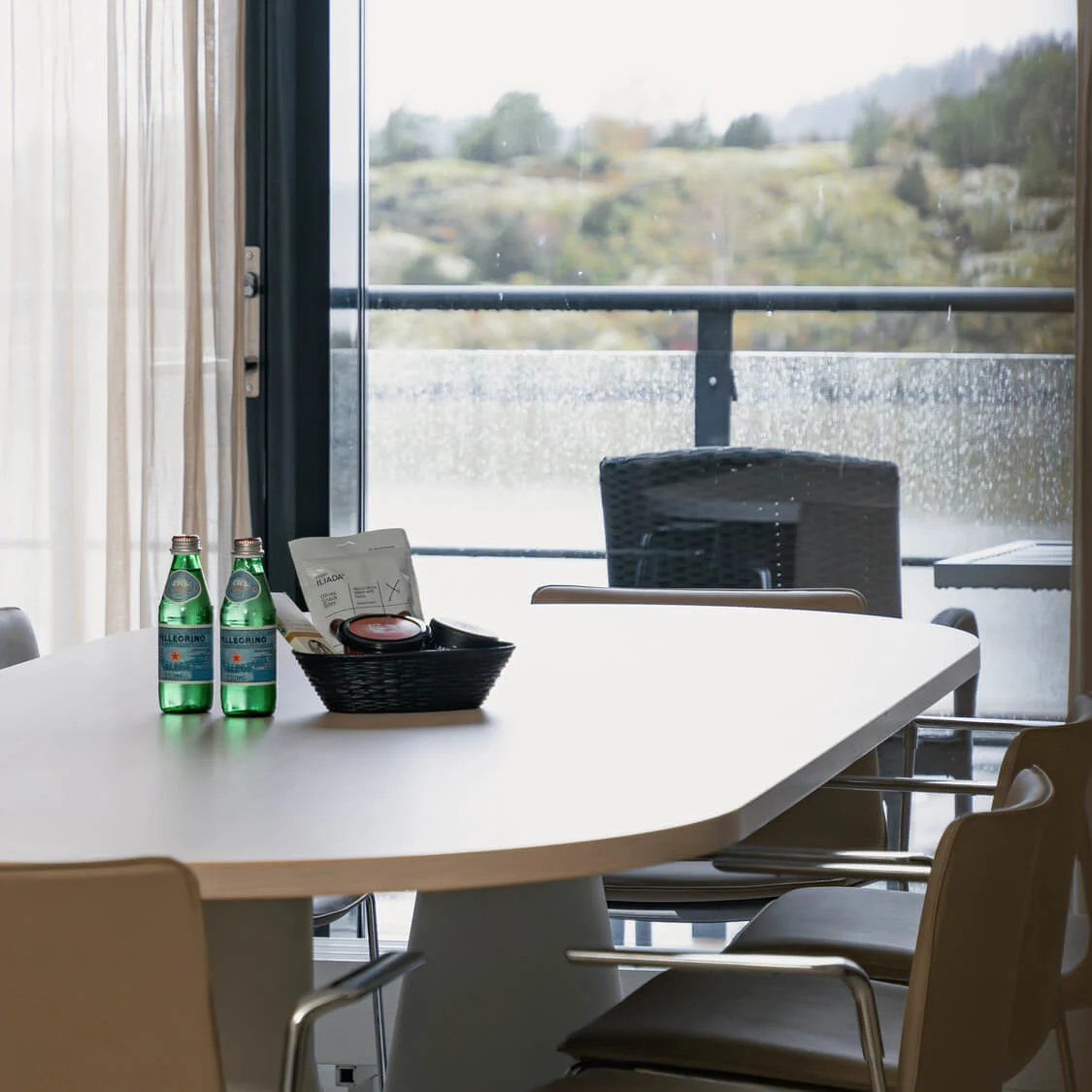 Et konferansebord har to flasker San Pellegrino og en kurv. Det er omgitt av stoler, med et stort vindu med utsikt over et regnvær.