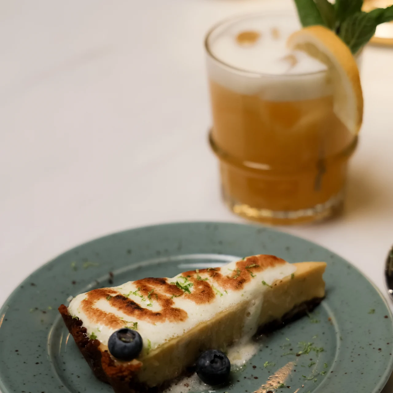 A slice of pie with a toasted top and blueberries sits on a green plate. Nearby, a frothy orange drink garnished with lemon and mint is on a table.