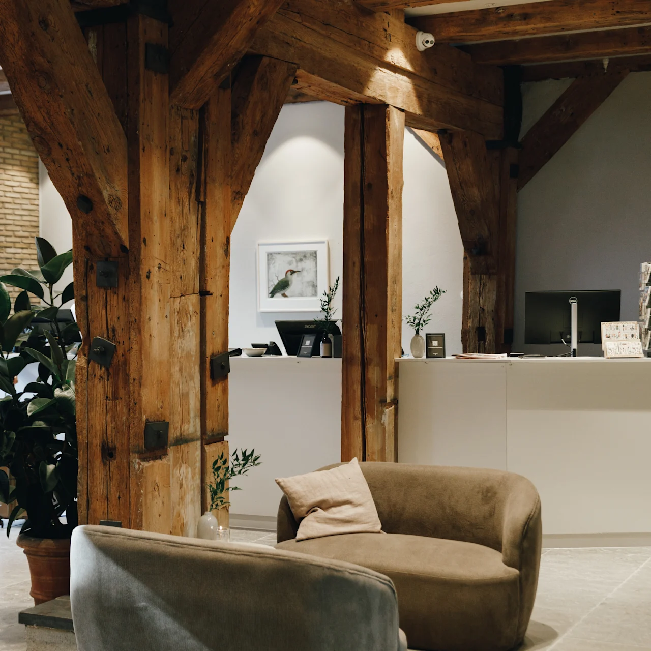 Beige and gray sofas are arranged around a wooden beam structure in a modern lobby with plants, a framed picture, and a reception desk holding computer monitors in a softly lit room.