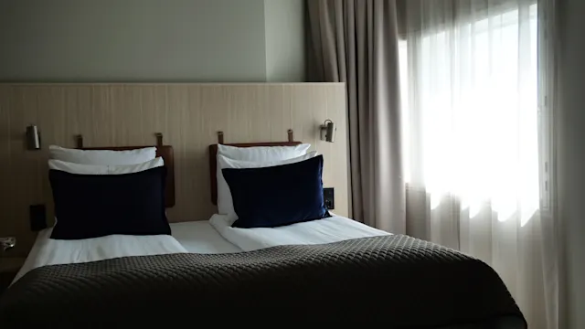 Siiististi pedattu sänky tummilla tyynyillä ja kuvioidulla peitolla, sijaitsee hämärästi valaistussa hotellihuoneessa, verhoilla varustetun ikkunan vieressä, josta tulee pehmeää luonnonvaloa.