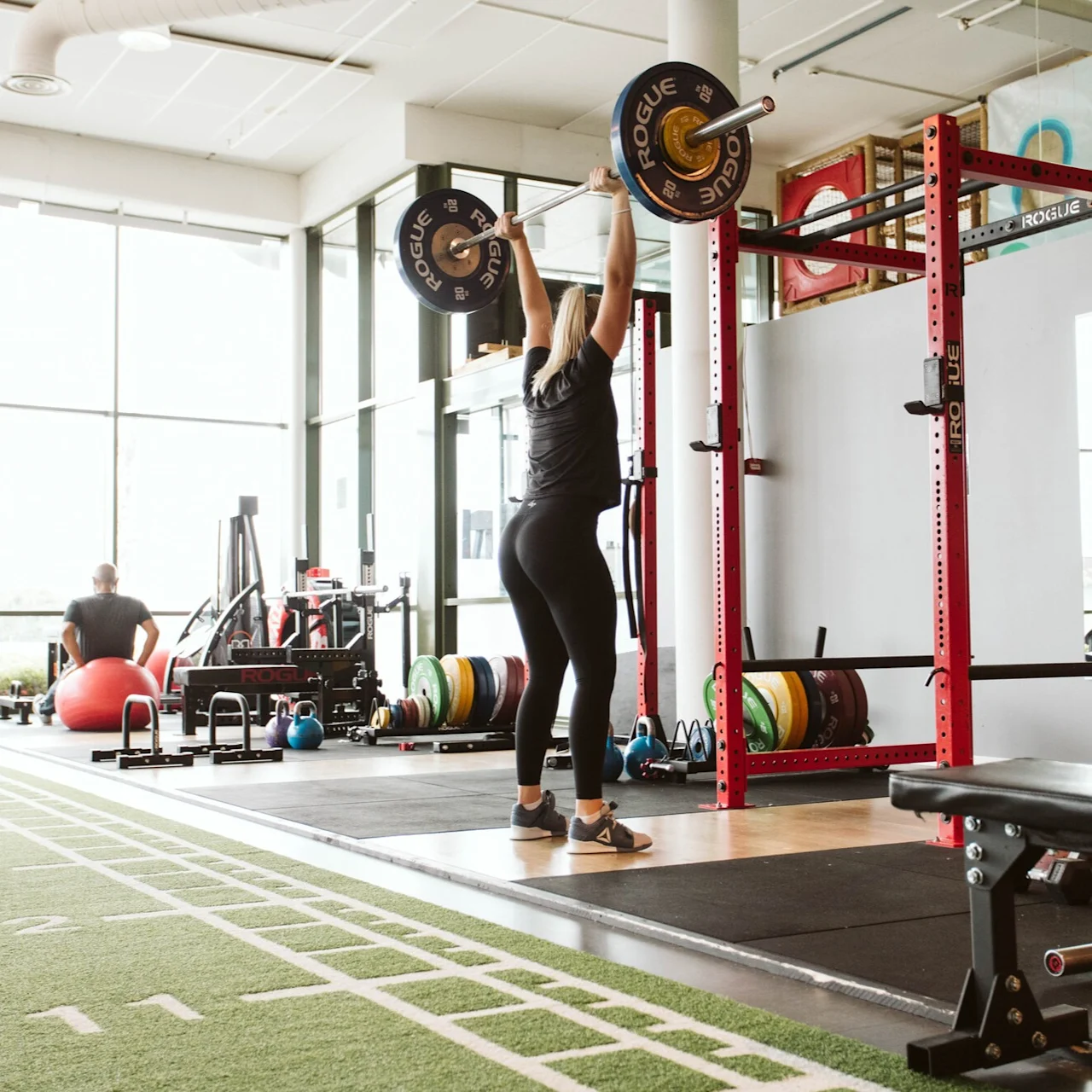 A person lifts a barbell overhead in a gym, surrounded by equipment, weight plates, and a green turf area. Visible text: "ROGUE."