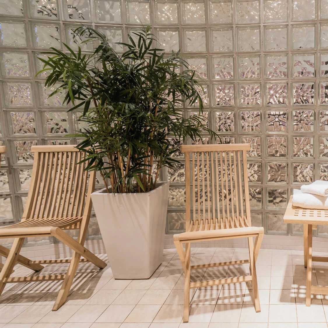 Wooden chairs and table with folded towels set in a tiled room; a potted plant is placed next to a glass block wall.
