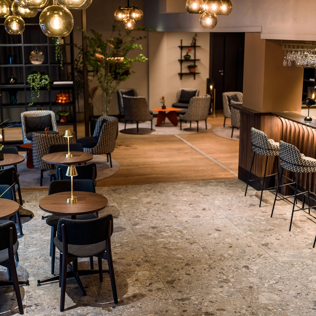 Cozy café interior; round tables with chairs, ambient overhead lighting, bar with stools, and plants create a welcoming atmosphere. Shelves and a small lounge area are visible in the background.