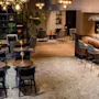 Cozy café interior; round tables with chairs, ambient overhead lighting, bar with stools, and plants create a welcoming atmosphere. Shelves and a small lounge area are visible in the background.