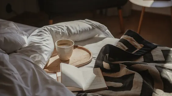 Viihtyisä sänkynäkymä, jossa on kahvikuppi, avoin kirja ja kuviollinen peitto, lämpimässä auringonpaisteessa.
