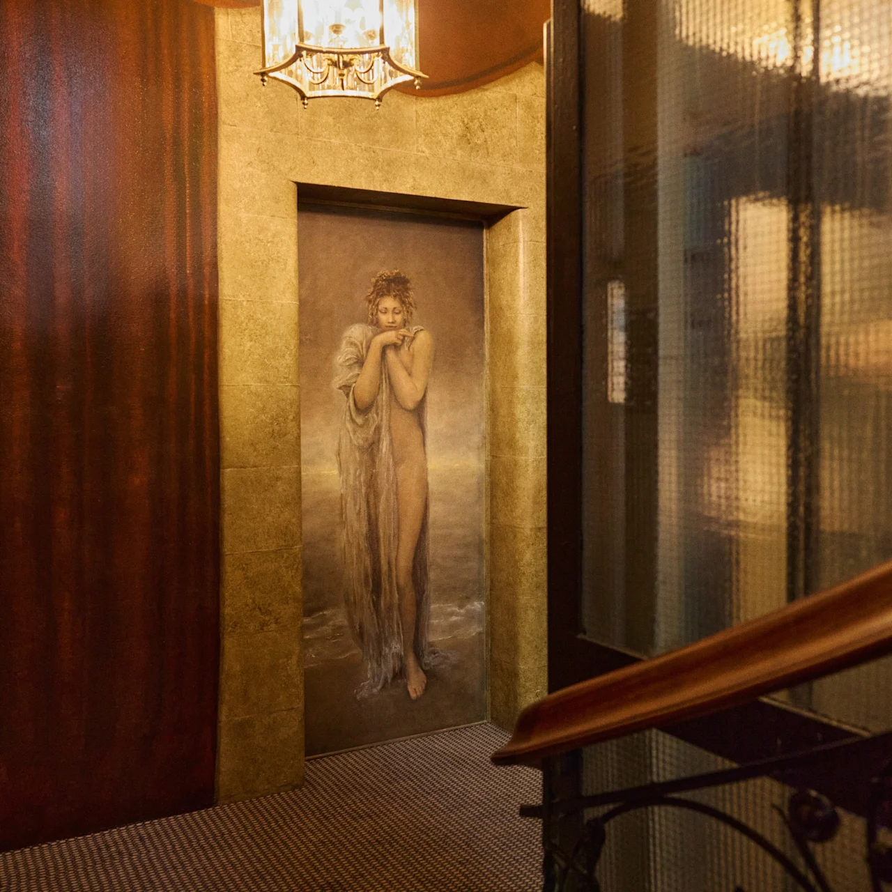 A painted door featuring a nude woman hugging a white angel stands in an ornate, warmly lit hallway with patterned walls and a metal railing on the right.