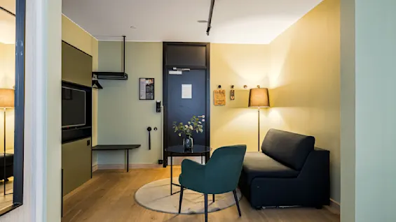Et koselig rom med en svart sofa, en grønn stol og et rundt bord med en plante. En gulvlampe gir mykt lys og skaper en varm atmosfære. Minimalistisk dekor og en dør med notater gir sjarm.