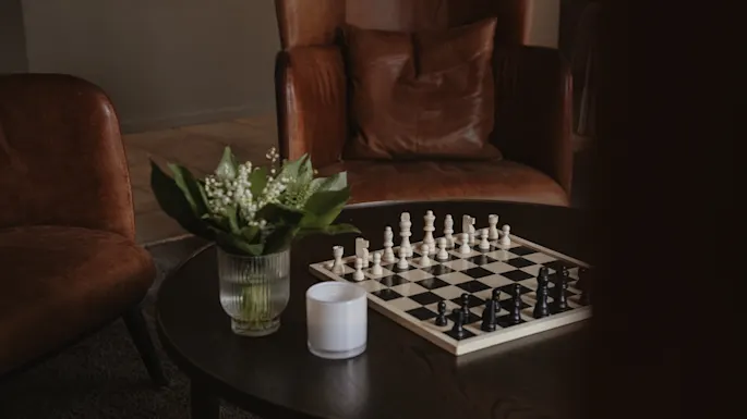 Et sjakkbrett med plasserte brikker står på et trebord, ved siden av en glassvase med hvite blomster, i et koselig rom med skinnstoler.