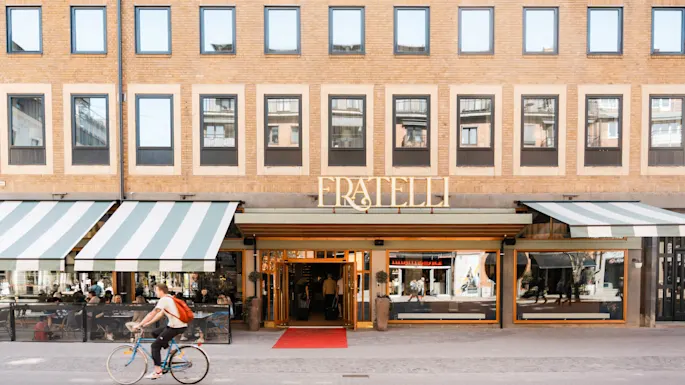 En syklist sykler forbi inngangen til en bygning med skiltet "FRATELLI" over, store vinduer og uteservering under stripete markiser på en solfylt dag.
