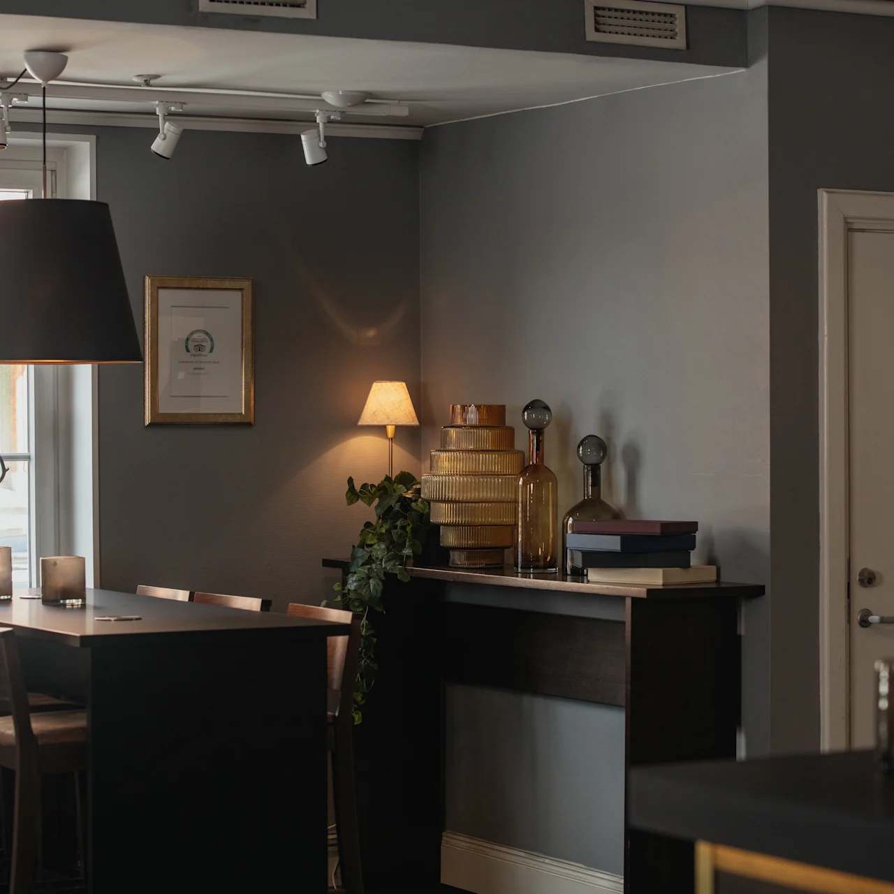 A dimly lit dining area features a tall black table with chairs, surrounded by gray walls. A lamp hangs overhead, and a door labeled "Damer" indicates a restroom.