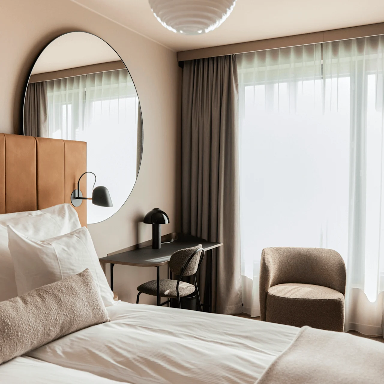 Bed with white sheets and pillows rests against a brown headboard. A round mirror, desk, and chair are adjacent. Large window with curtains brightens the room.