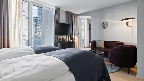 A hotel room features two neatly made beds, a wall-mounted TV, and a maroon sofa beneath a floor lamp, with cityscape views visible through large windows.