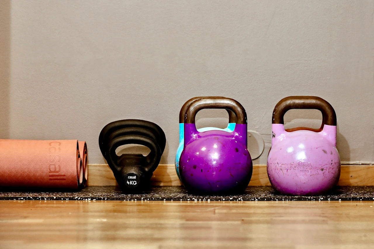 A black kettlebell labeled "4 KG" sits between two larger kettlebells. An orange foam roller lies parallel nearby, all on a wooden floor against a beige wall.