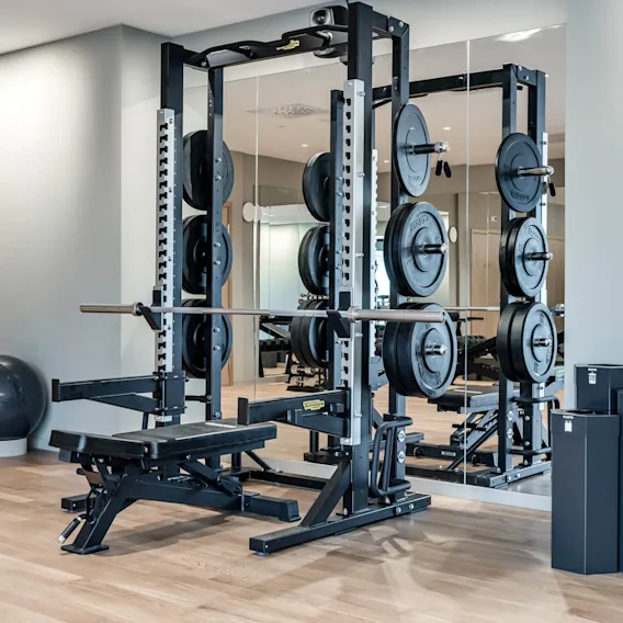 A weightlifting bench with a loaded barbell stands in a gym room. Mirrors, exercise balls, and storage boxes define the sleek, minimalist environment.