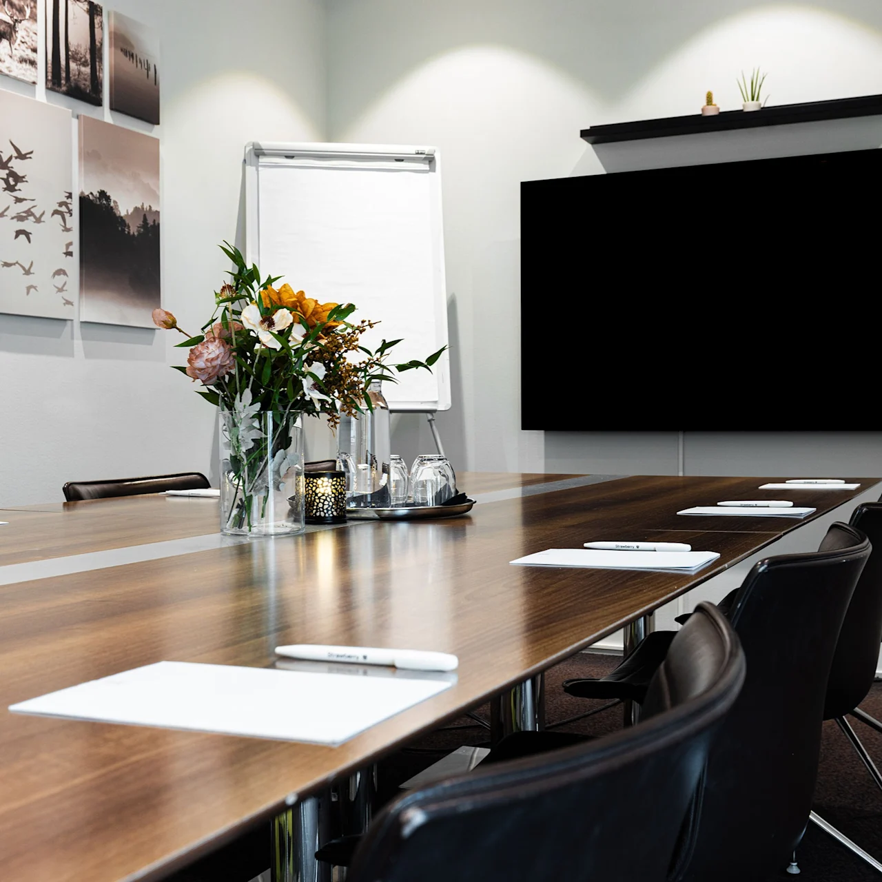 A long wooden table is set for a meeting with chairs, notepads, and pens; a bouquet and flip chart are central. Walls display art and a large screen.
