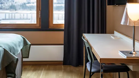 A desk with a lamp and chair is placed against a wall beside a large window, overlooking a snowy landscape with a bed positioned near the window.