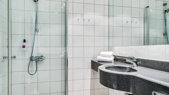 A glass shower with a chrome showerhead is adjacent to a dark stone sink in a white-tiled bathroom. Towels are neatly stacked on a shelf.