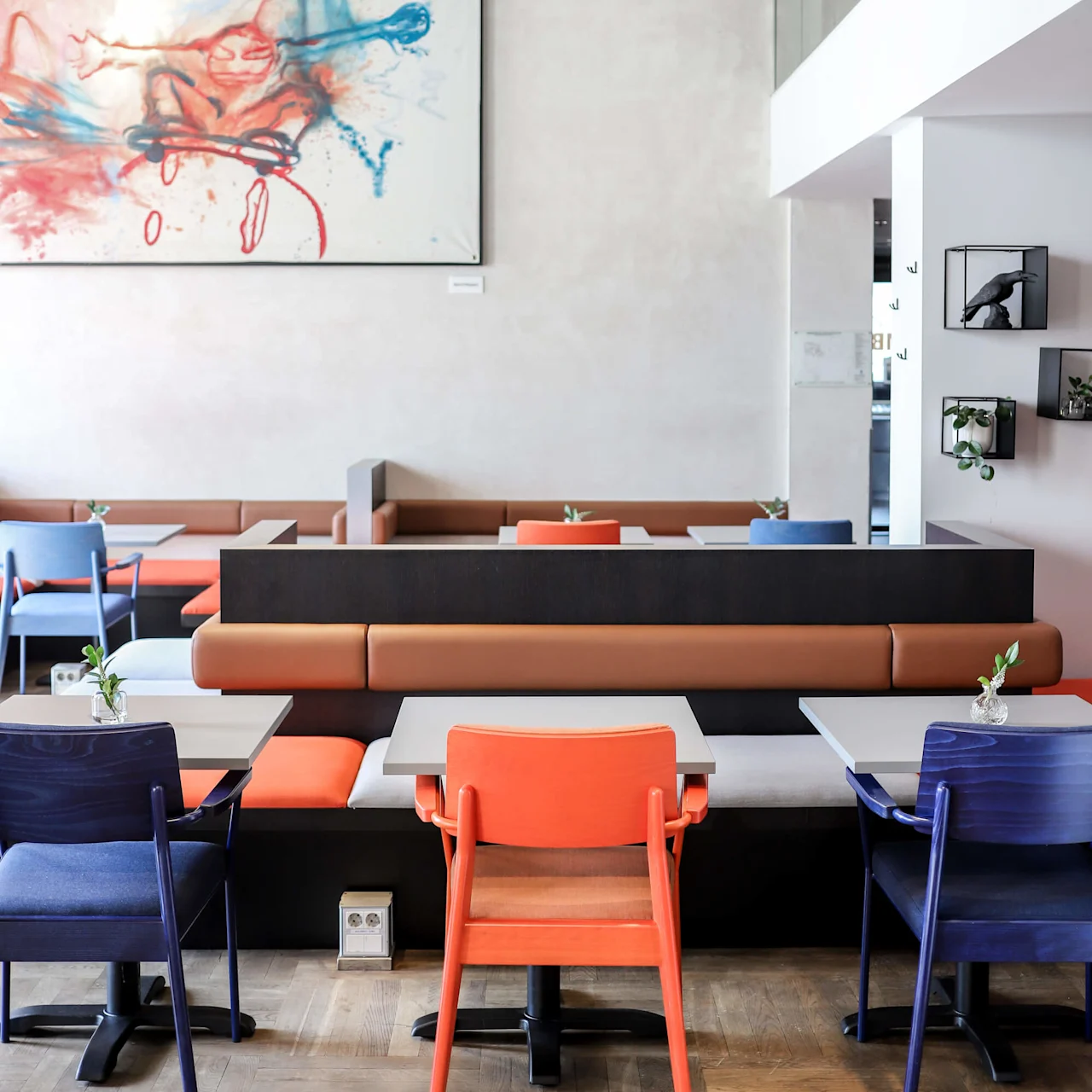 Dining tables with colorful chairs are arranged in a modern café. Abstract art decorates the wall, with potted plants and warm lighting enhancing the cozy atmosphere. 