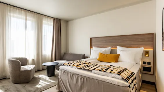 Valoisa ja moderni hotellihuone, jossa on mukava sänky, viihtyisä nojatuoli ja suuret ikkunat.