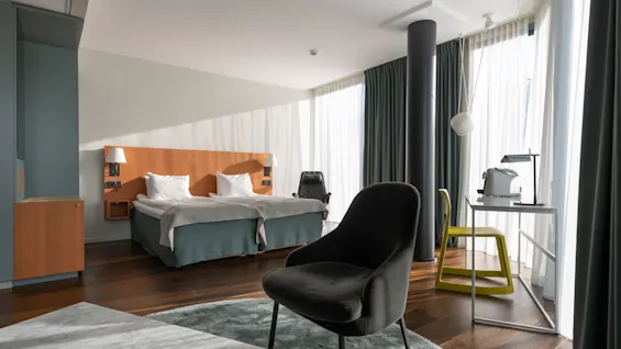 Et moderne hotellrom med en dobbeltseng, hvite puter, en svart lenestol og et skrivebord. Gulv-til-tak-gardiner slipper inn sollys og skaper en lys atmosfære.