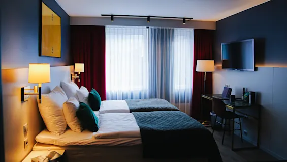 Ett modernt hotellrum med två sängar med vita och gröna sängkläder, belysta av vägglampor. Ett skrivbord med en bärbar dator står under en väggmonterad TV, nära stora fönster med gardiner.