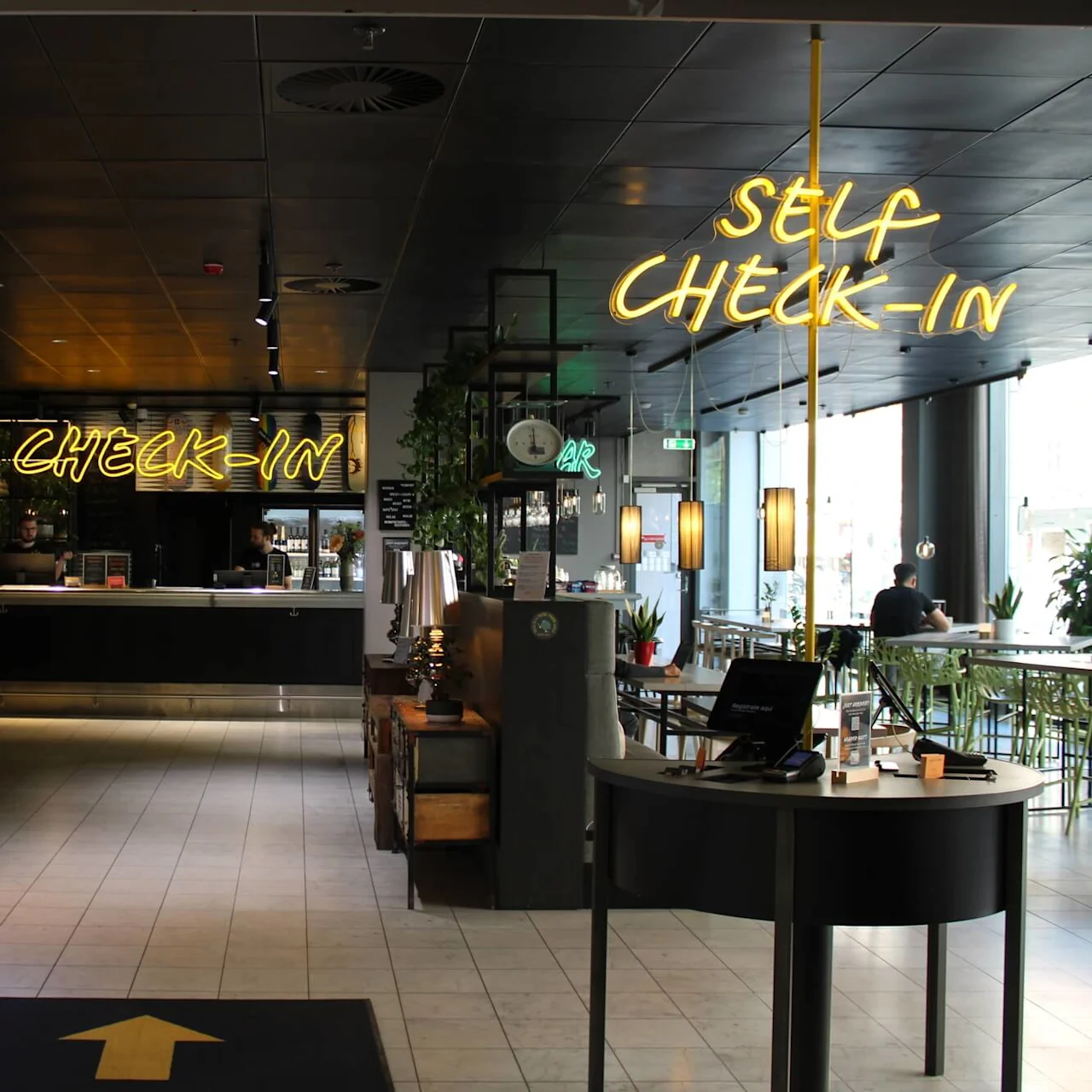A modern hotel reception area with a self-service check-in station. Neon signs on the wall display the words "CHECK-IN" and "SELF CHECK-IN".