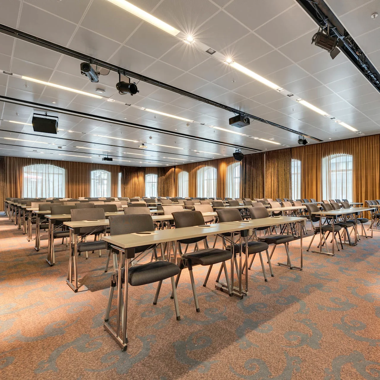 Rækker af tomme stole vender mod en scene med et podium i et rummeligt konferencelokale, der har mønstret tæppe, store vinduer med gardiner og loftsbelysning.