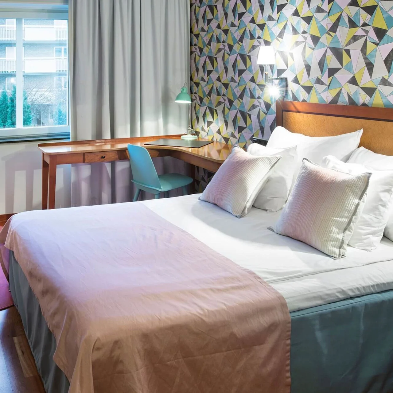 A bed with pillows rests in a room. A patterned wall, a desk with a lamp, a blue chair, and a beige armchair on a pink rug complement the cozy setting.