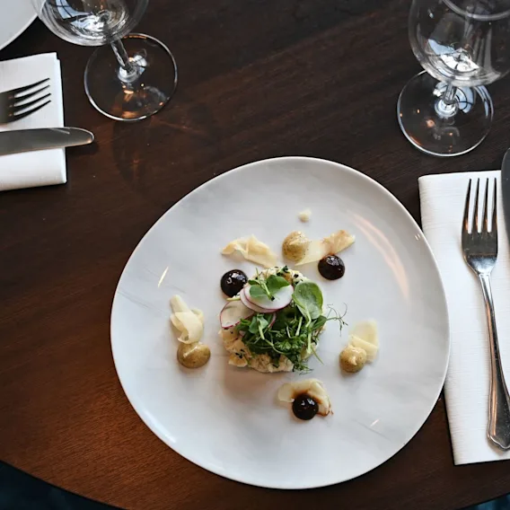 A plated dish of assorted greens, radish slices, and sauces, is elegantly arranged on a white plate. Wine glasses and cutlery rest on a dark wooden table.