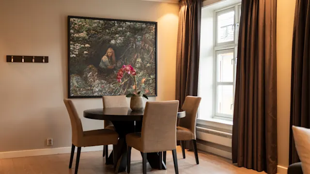 Et rundt spisebord med fire beige stoler omgir en vase med rosa blomster. Et utsmykket maleri henger på en beige vegg, og et stort vindu med mørke gardiner er i nærheten.