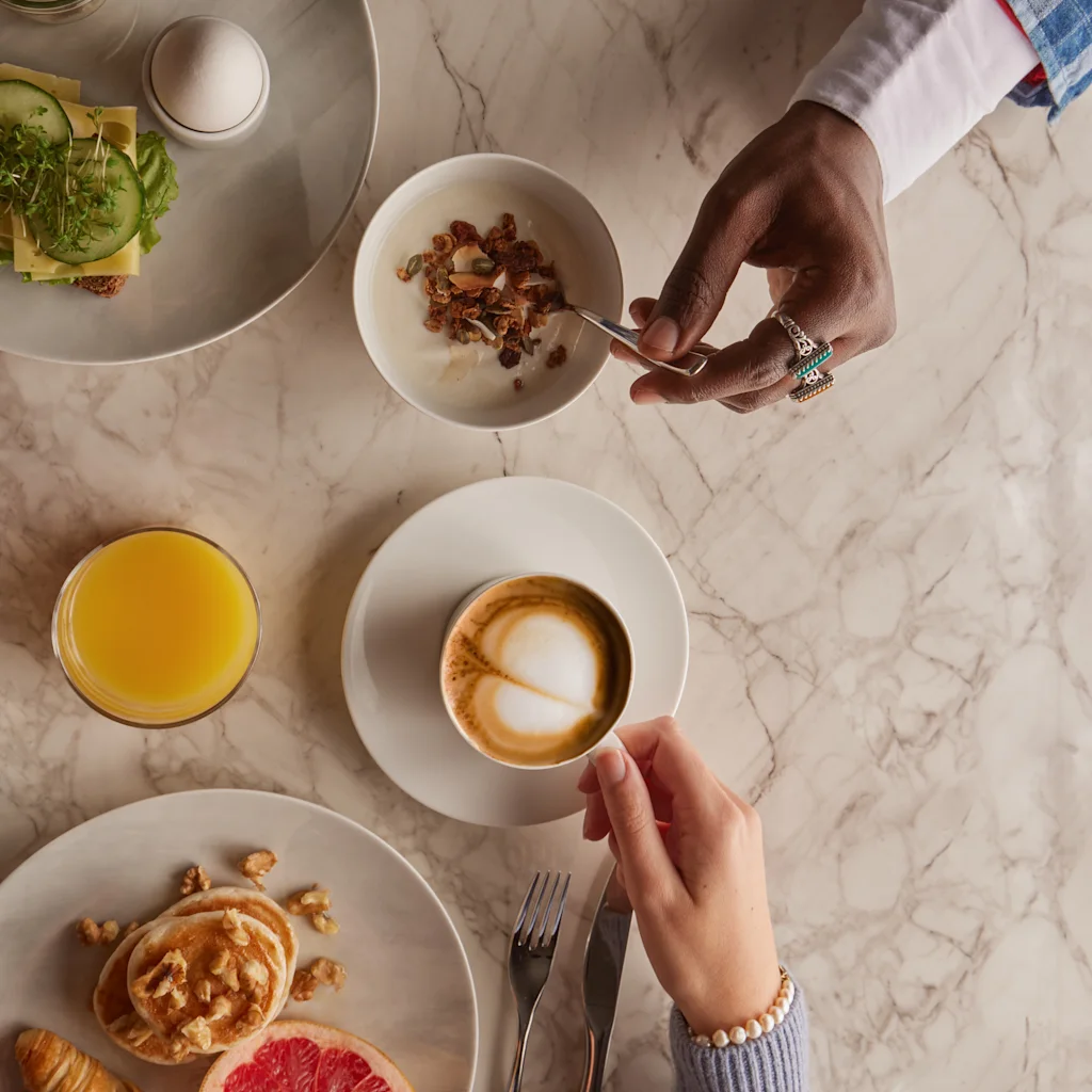 Et bord viser frokostmat: en hånd heller granola i yoghurt, en annen holder en cappuccino. I nærheten er toast med agurk, et kokt egg, appelsinjuice, en croissant og grapefrukt.