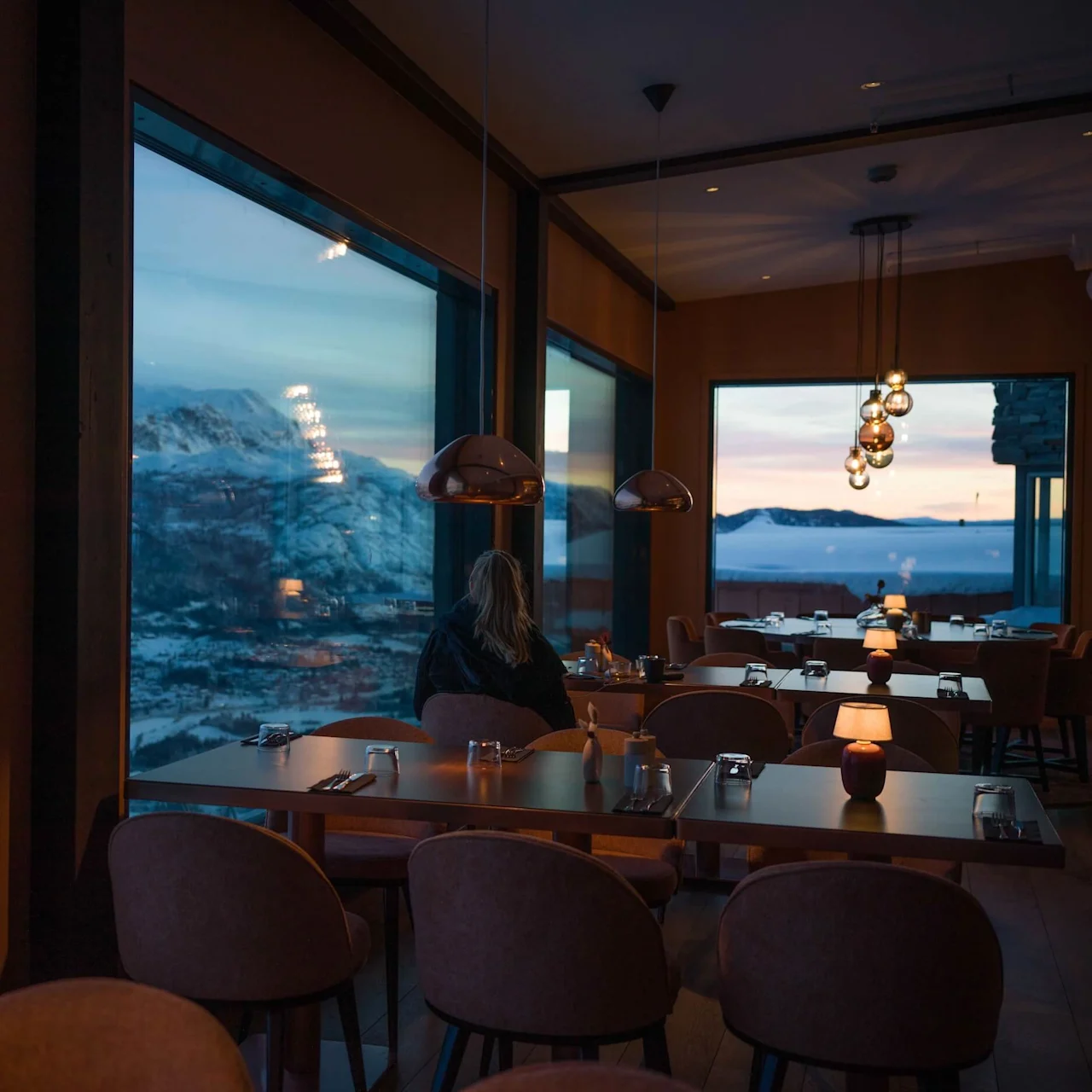 Large windows showcasing snow-covered mountains surround dimly lit restaurant tables, where a solitary person sits. Pendant lamps hang above, and soft candlelight enhances the cozy atmosphere.