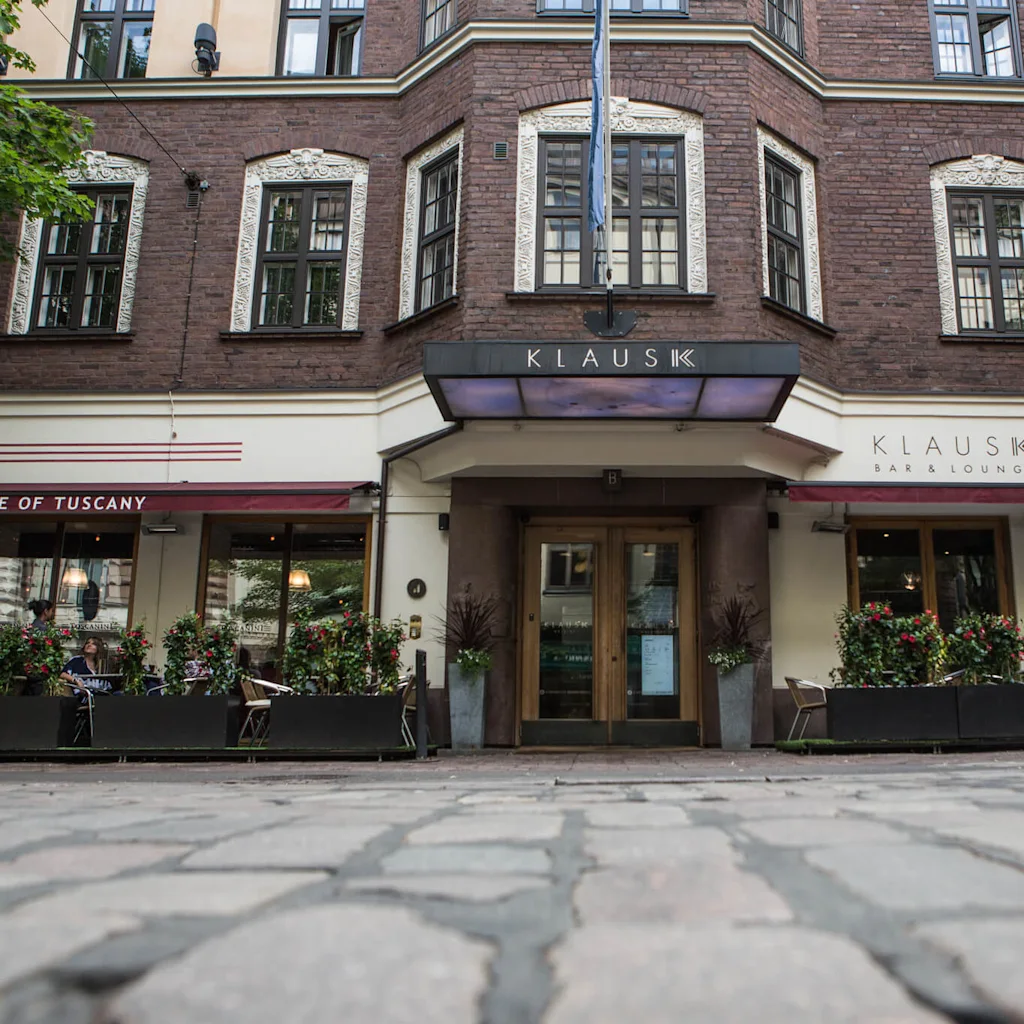 Byggnadsfasad med "KLAUS K"-skylt ovanför ingång. Gatumiljö med ett kafé under röd markis märkta "A TASTE OF TUSCANY" och uteservering omgärdad av växter.