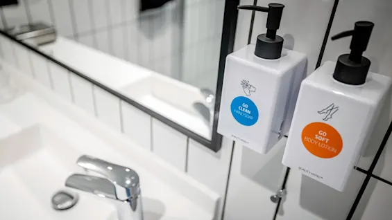 Tvåljus- och kroppslotiondispensrar monterade på en kaklad badrumsvägg, reflekterade i spegeln. Text: "GO CLEAN HAND SOAP" och "GO SOFT BODY LOTION," "QUALITY HOTEL™."