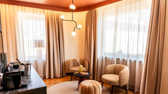 A cozy living area features two armchairs and a round ottoman on a carpet, with a table adorned with a vase and snacks, under a modern chandelier and surrounded by windows with curtains.