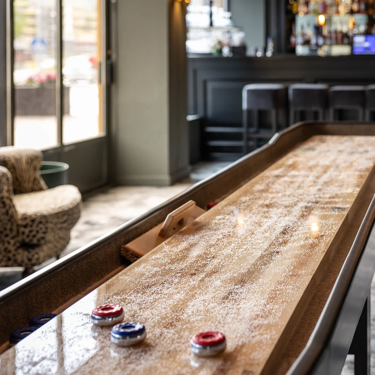 Shuffleboardbord med røde og blå pucker står i en dæmpet belyst bar med et mønstret gulv og en godt udstyret bardisk i baggrunden.