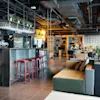 A modern bar area with stools and various seating arrangements in a spacious industrial-style lounge with exposed ductwork and warm lighting.