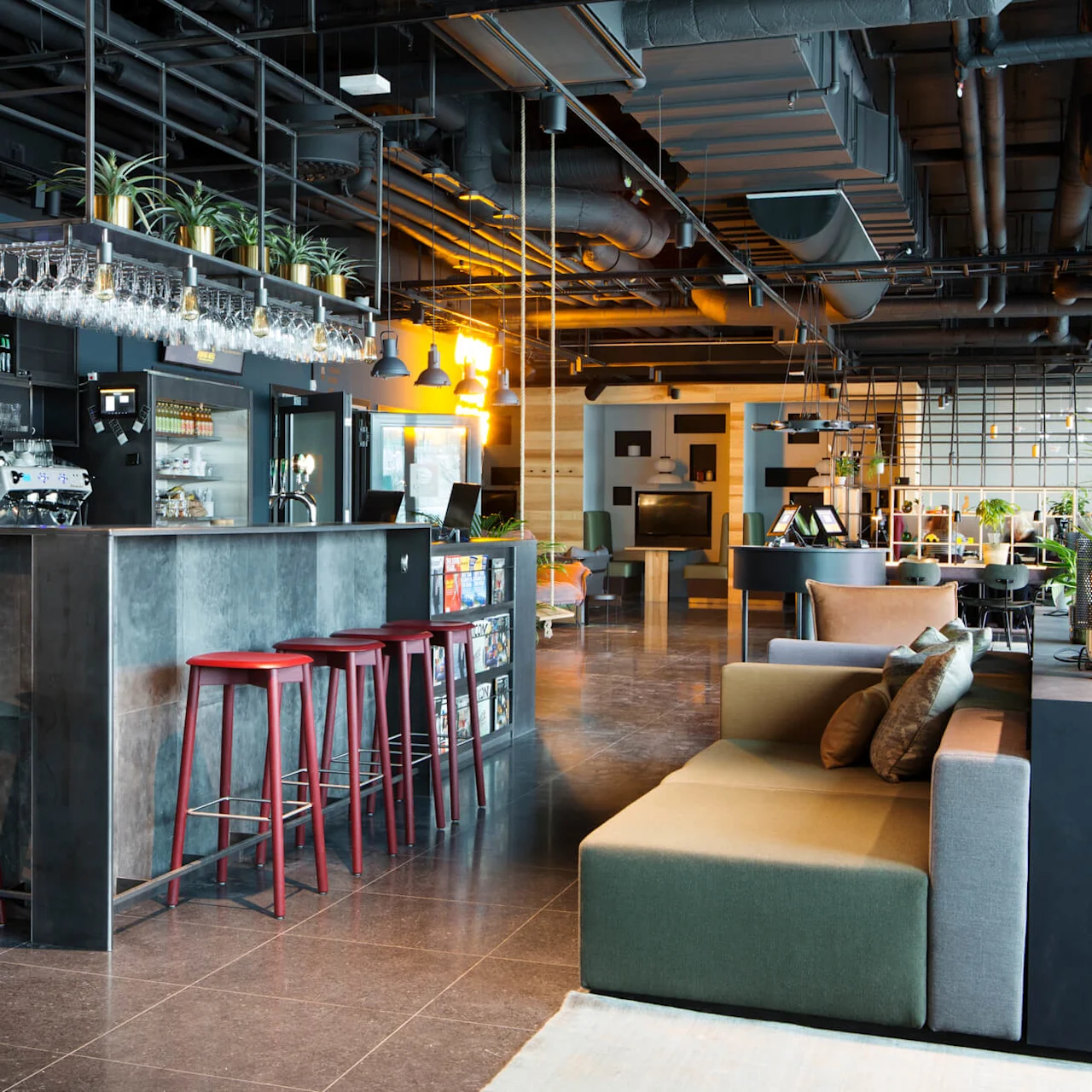 En modern bar med pallar och diverse sittgrupper i en rymlig lounge med industriell stil, synliga ventilationsrör och varm belysning.